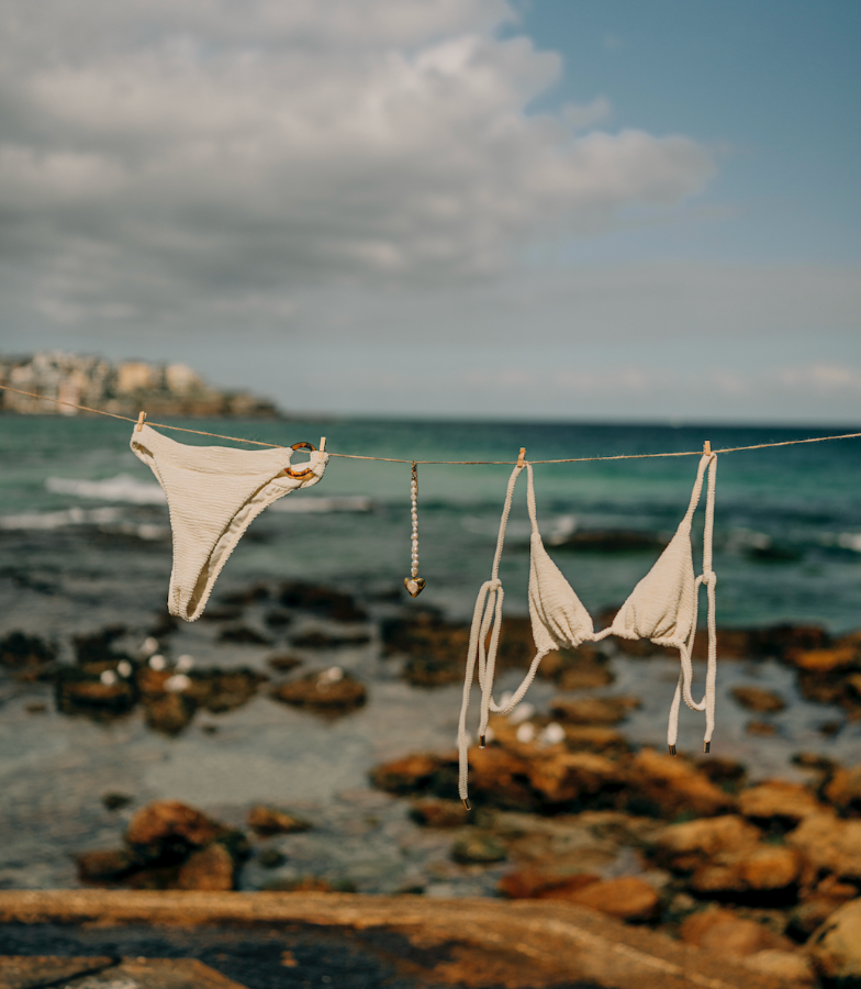 Beige bikini hanging on a clothesline by the ocean with rocky shoreline and cloudy sky.