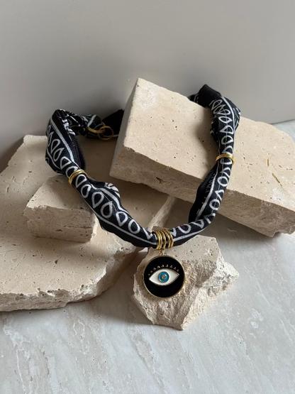 A black and gold colored necklace with a paisley bandana pattern and a gold stainless steel charm, displayed on a beige stone surface.