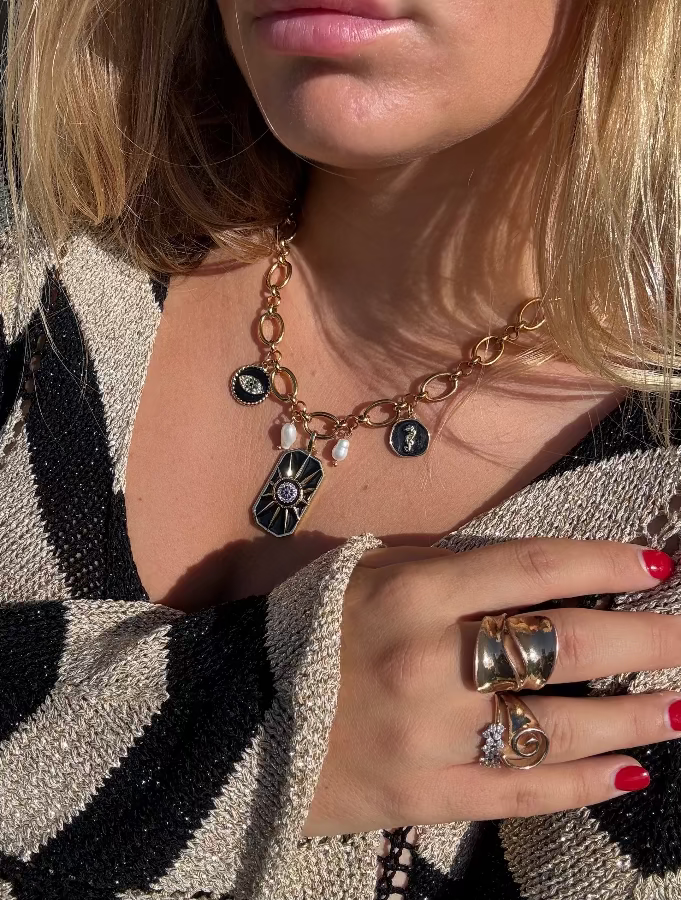 Close-up of a person wearing a gold necklace with charms and multiple rings on a patterned fabric background.