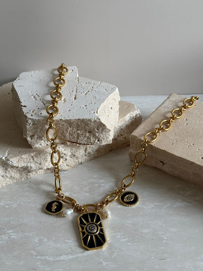 A gold stainless steel necklace with charms and freshwater pearls displayed on a stone surface.
