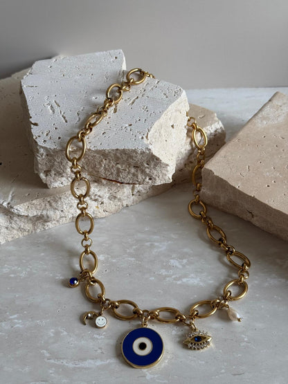 A gold-colored stainless steel necklace with charms and a freshwater pearl, displayed against a neutral-toned textured background.
