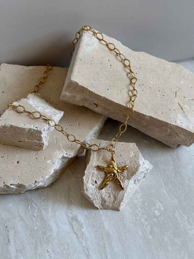 A gold stainless steel necklace with a star charm, placed on a textured white surface.