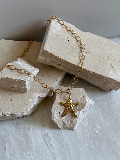 A gold stainless steel necklace with a star charm, placed on a textured white surface.