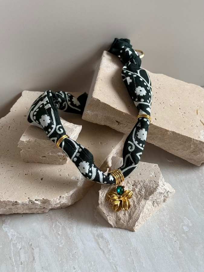 A black and white paisley bandana style necklace with a gold stainless steel charm, displayed on a beige stone surface.